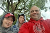 A selfie of two boys wearing hoods and a man with a tree in the background