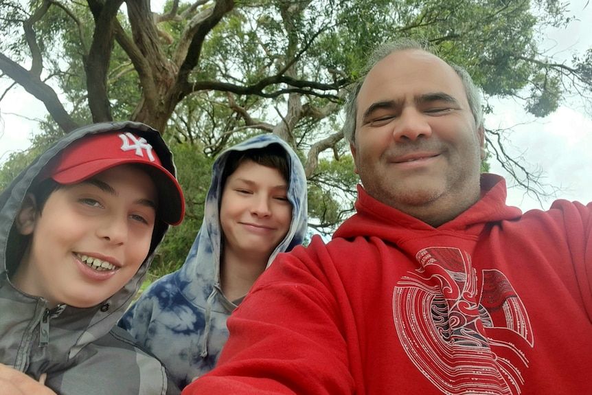 A selfie of two boys wearing hoods and a man with a tree in the background