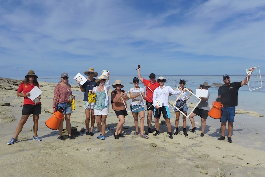 11 people stand on a white sand beach, clear water, blue sky behind.