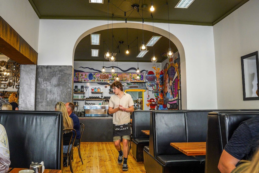 A boy in a shirt and half-apron approaches a table inside a tall-ceiling cafe space with booths.