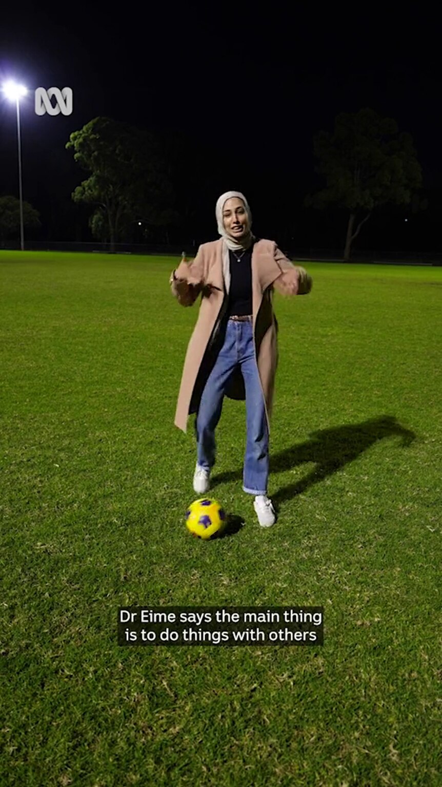 A young woman with medium-tone skin in a hijab and jeans stands with a soccer ball on grass at night
