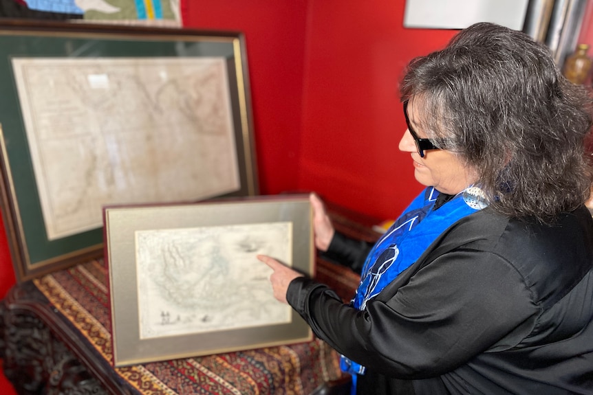 A woman with dark hair sits at a desk inside, pointing at a framed historical map.