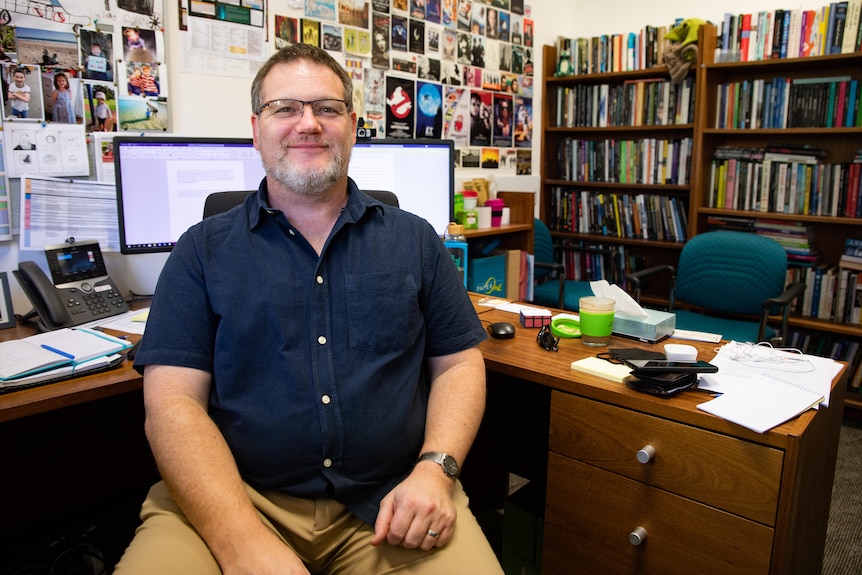 A man sitting at his desk with books and posters behind.