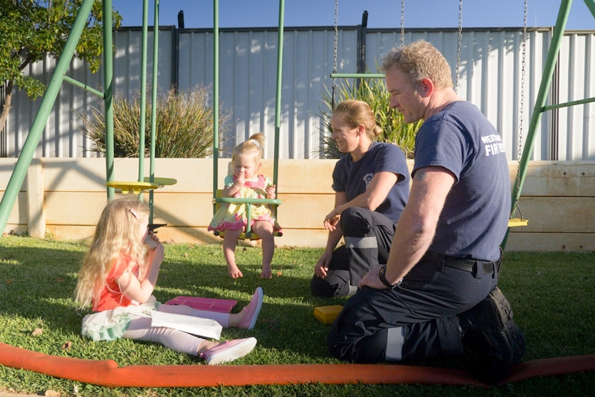 A husband and wife in fire brigade t-shirts, kneel on the grass by a swing set with two little girls playing in a backyard.