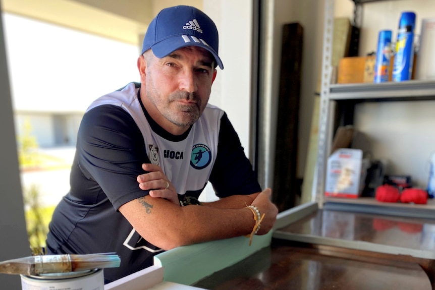 Man standing in a garage with cap on, looking at the camera while leaning on an old piece of furniture with some paint on it.