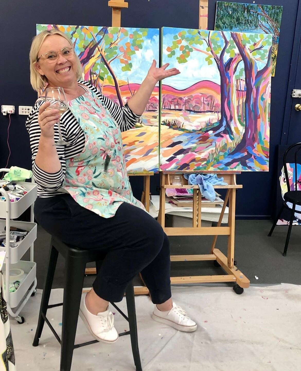 a women sitting with a glass of wine, a colourful landscape painting behind her