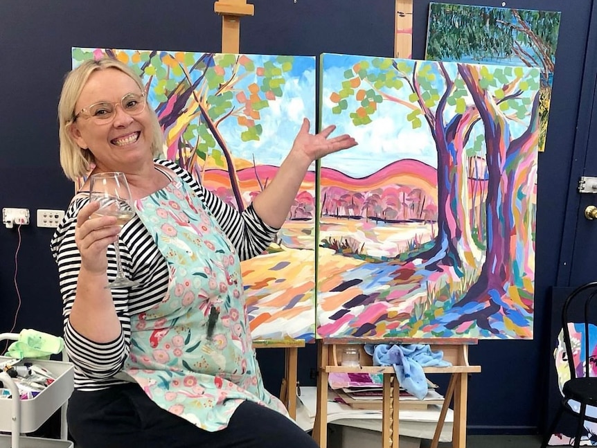 a women sitting with a glass of wine, a colourful landscape painting behind her