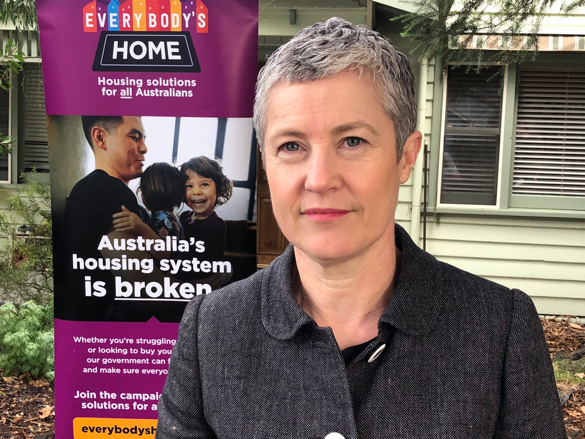 A woman with short grey hair and a dark grey jacket stands in front of a banner saying "Australia's housing system is broken".
