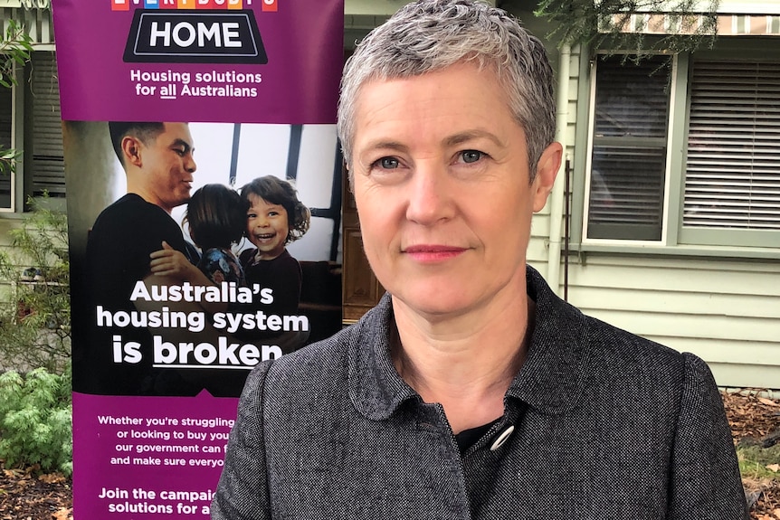 A woman with short grey hair and a dark grey jacket stands in front of a banner saying "Australia's housing system is broken".
