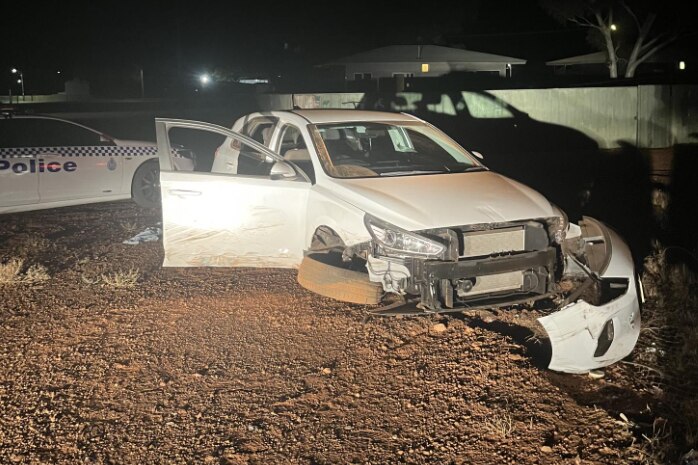white car at night in the dark with front part ripped off, police car in the background 