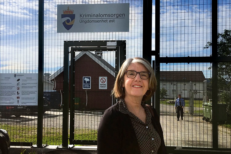 An older woman stands in front of large security gates with red building and a security officer in background.