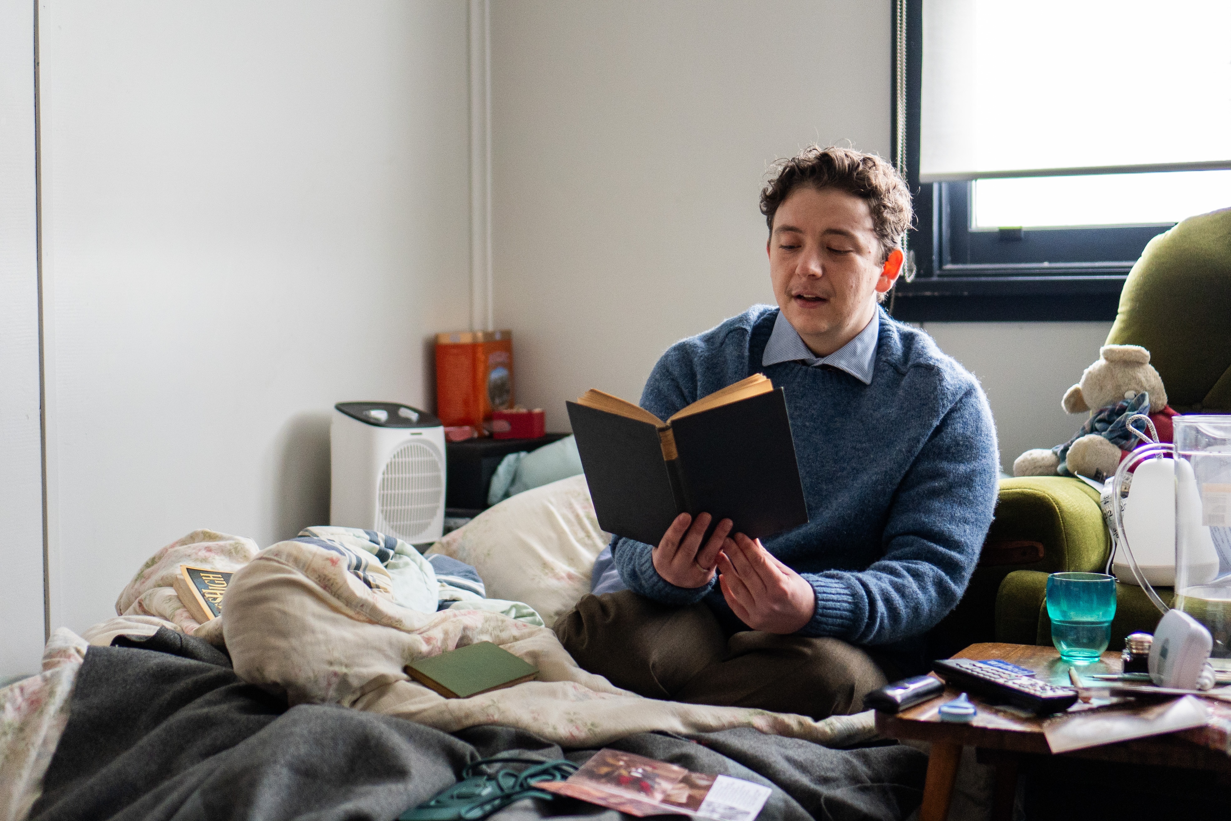A young man sitting on a mattress, reading aloud from an old-looking book.