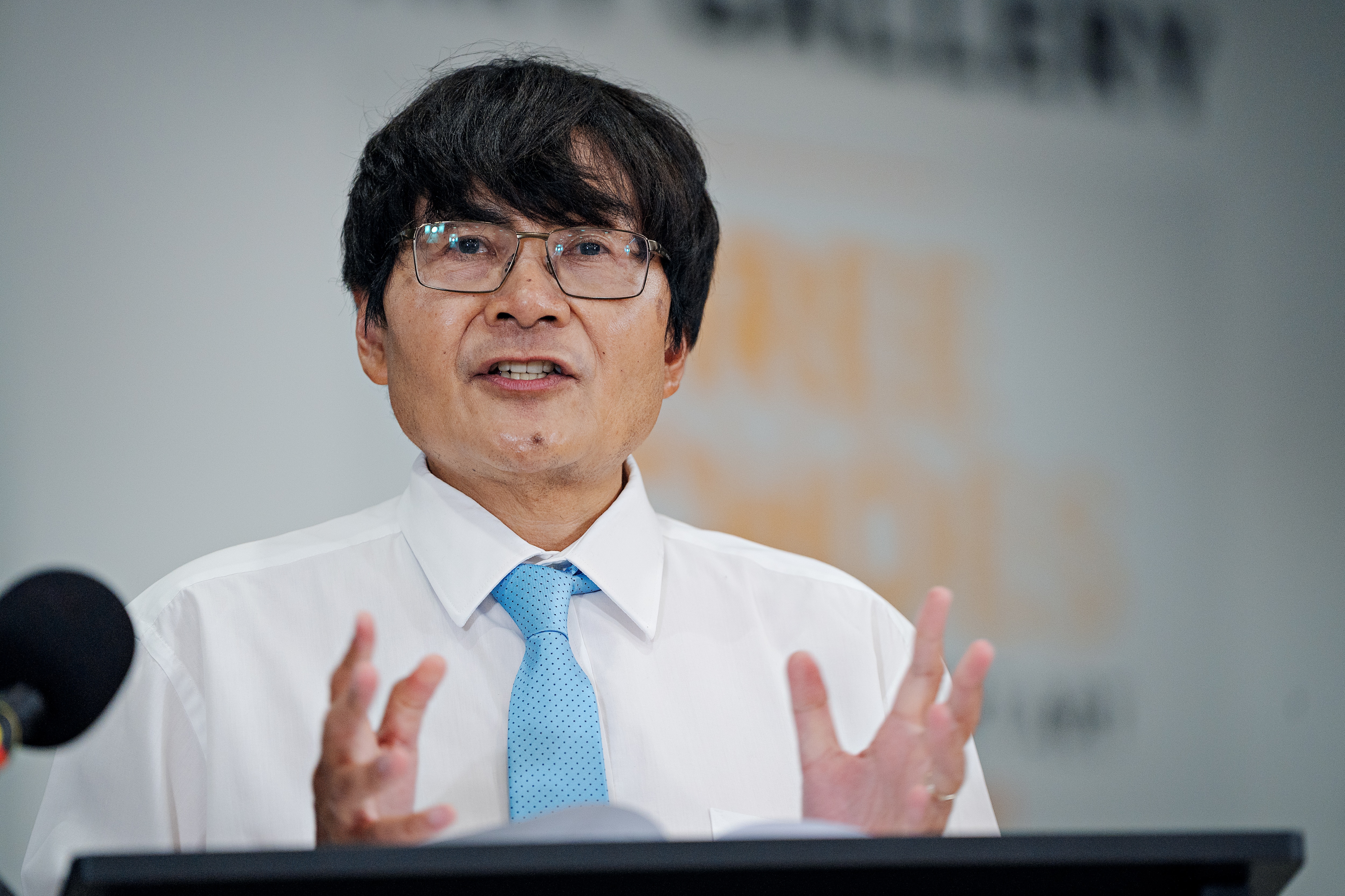 Um homem com óculos de leitura e gravata azul, discursando em uma conferência.