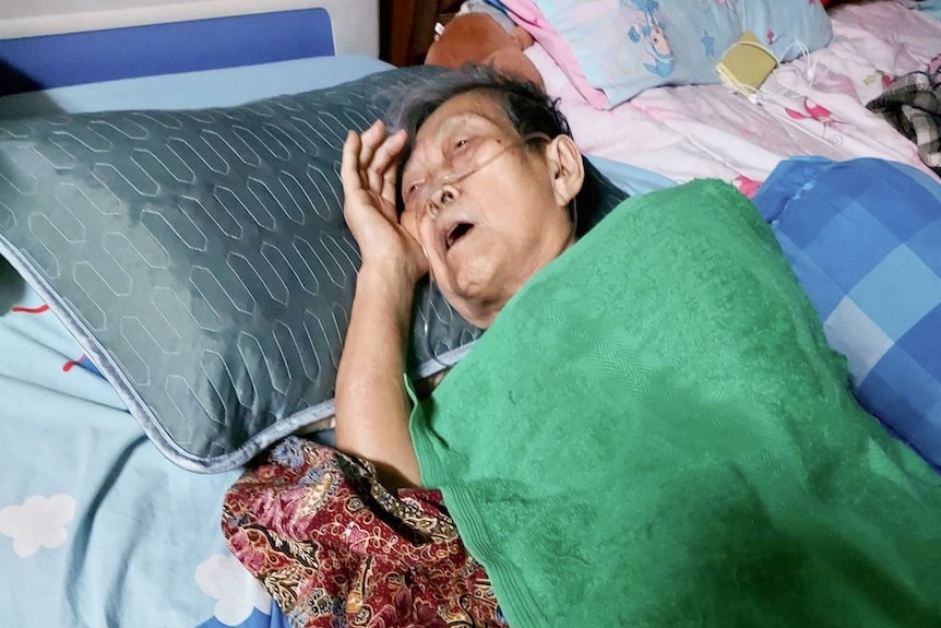 An older Thai woman in a mask lies on a bed 