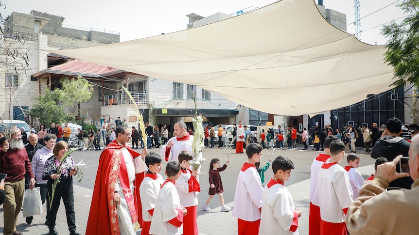 children, adults, and priests, some holding palm leaves, walking in a circle amongst white buildings 