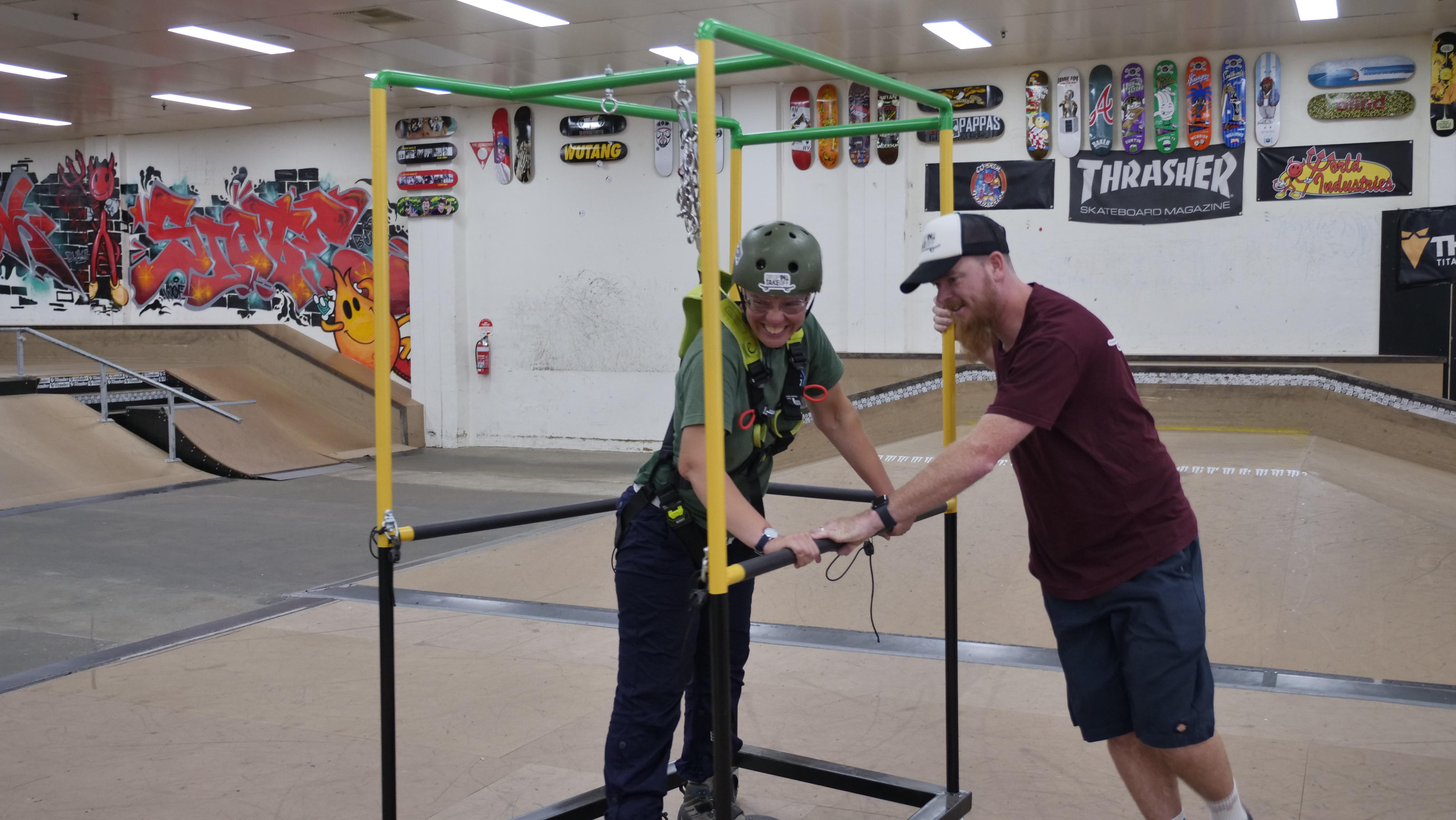 A man pushes a rectangular prism-shaped skate frame, while the woman on a skateboard inside the frame laughs