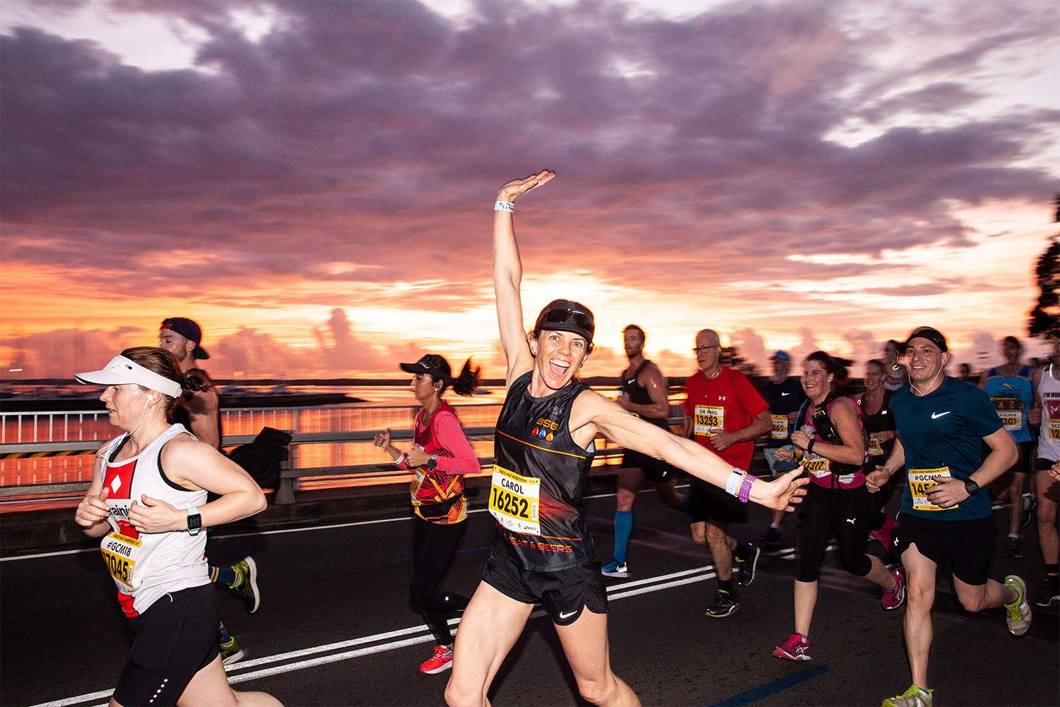 A woman in black running clothes with a group of runners, smiles to the camera and stretches out her arms at sunrise.