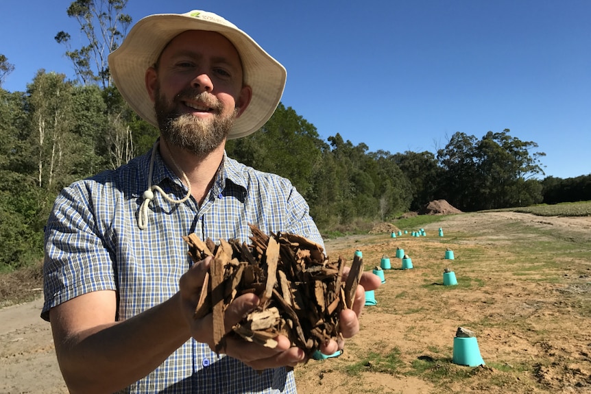 A man with a beard and a floppy hat stands in front of upturned buckets with woodchips in his hands