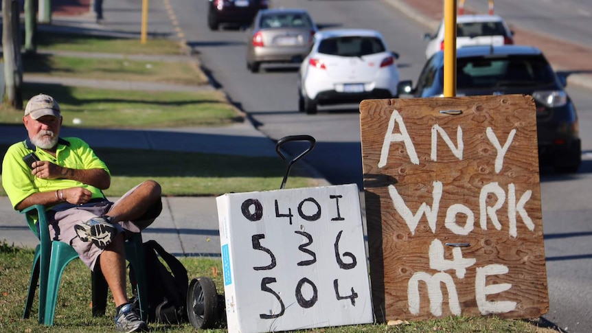 Les Devlin sitting on a plastic chair by a busy road with a sign reading 'Any work 4 me'.