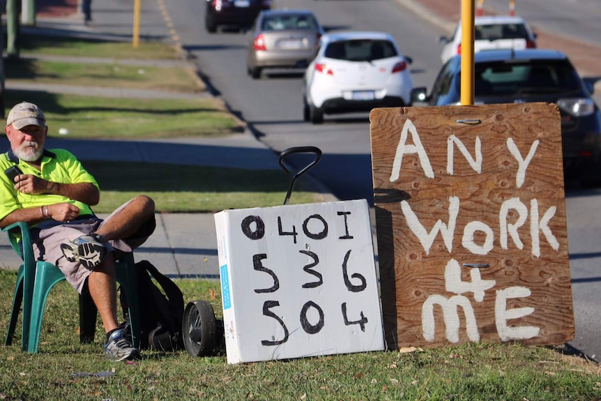 Les Devlin sitting on a plastic chair by a busy road with a sign reading 'Any work 4 me'.