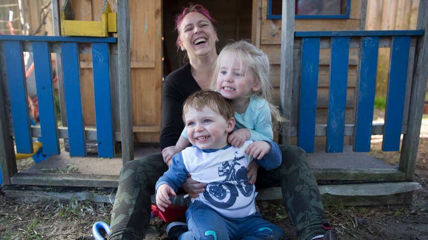 A mother and her two children sit in front of a cubby house.