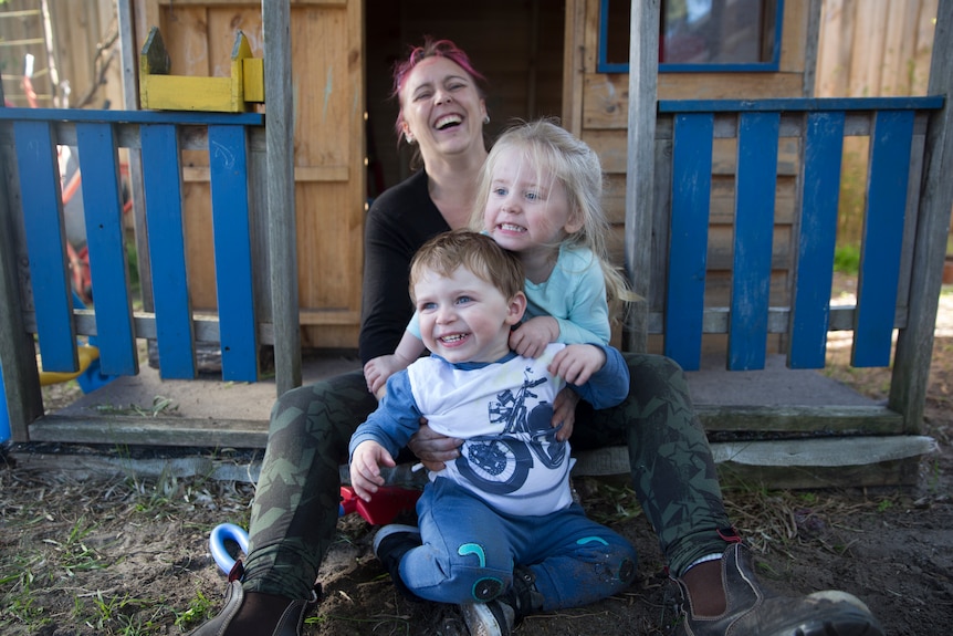 Amber, Nara and Roman sit in front of a cubby house.