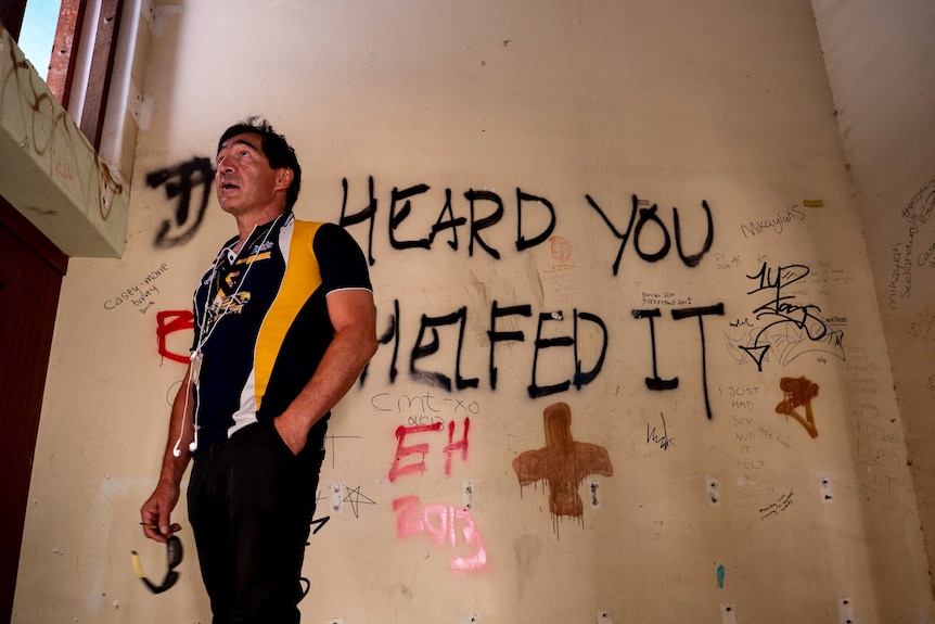 Man stands at the top of a staircase. The wall behind is covered in graffiti, he looks up towards the roof looking concerned.