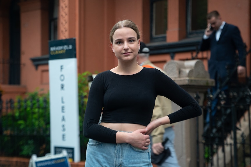 a young woman with blonde hair wearing a black long sleeved shirt and jeans stands in front of a for lease sign.