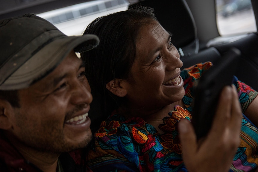 A man and woman with big smiles sit in the back seat of a car looking out the window 