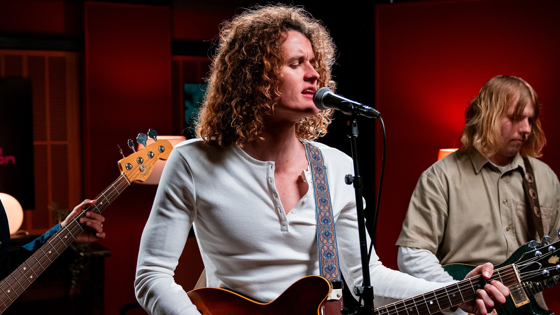 A curly-haired man in a white longsleeve sings into a microphone and plays guitar. Another man plays guitar behind him