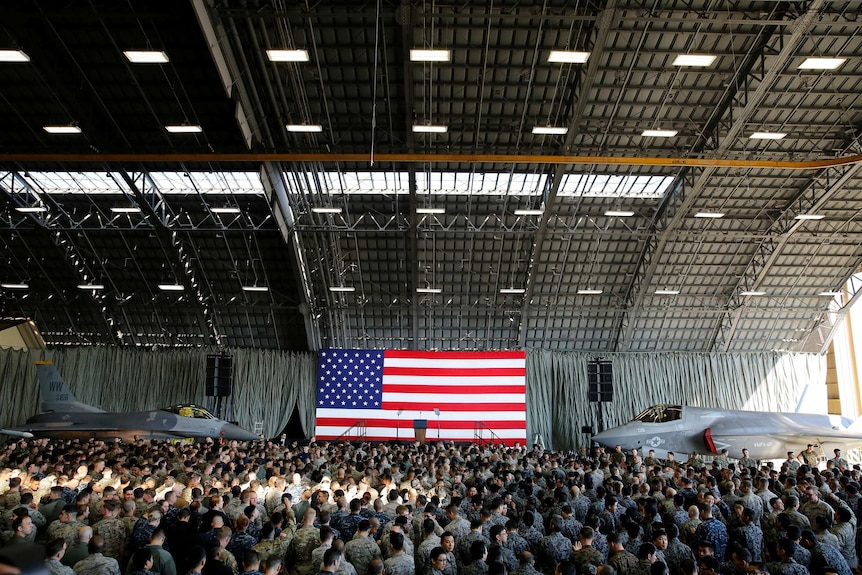 A hangar, with a large American flag behind an empty podium, is filled with people in military garb and two fighter planes.