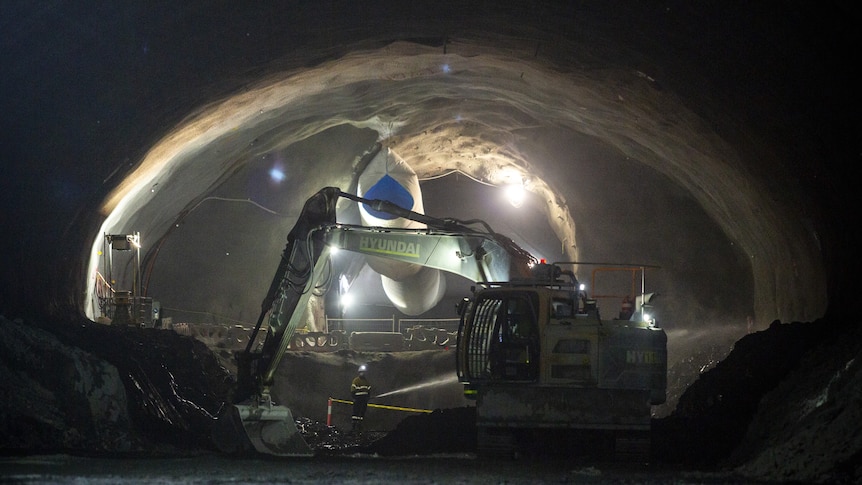 A worker waters the ground in the distance while in a tunnel and a excavator works alongside them