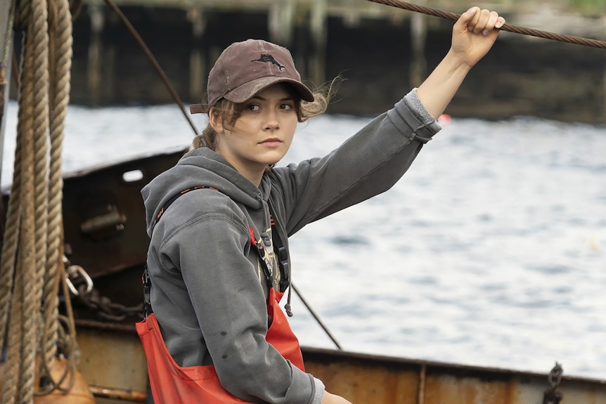 A teenage girl sits in a fishing boat looking glum, wearing a sweatshirt and cap