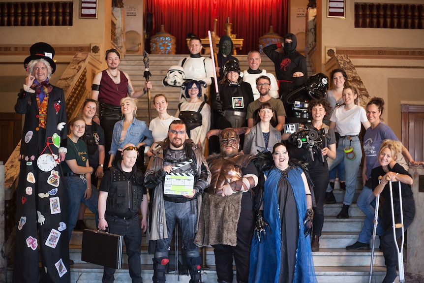 24 people stand on the steps of a theatre. Some wear costumes and others are in regular clothes. One woman holds a video camera.