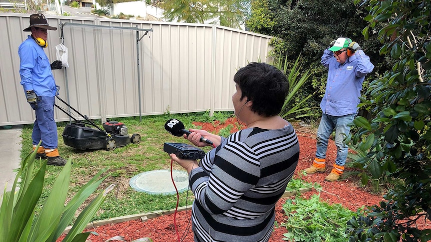 A woman holding a microphone records the sound of two men mowing the lawn.