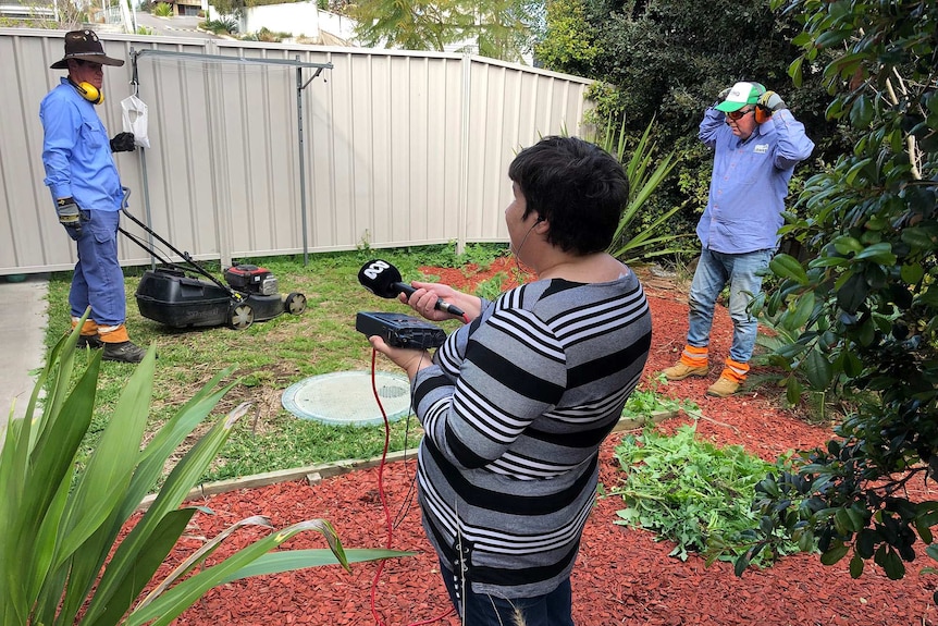 A woman holding a microphone records the sound of two men mowing the lawn.