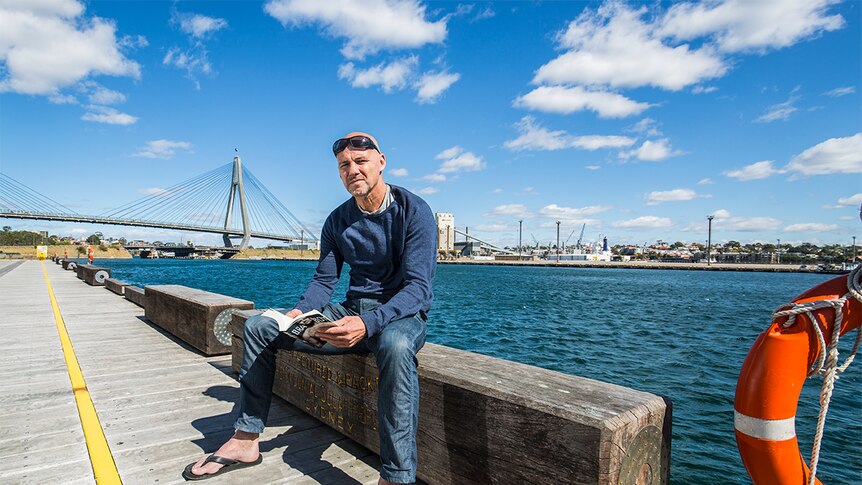 Gary Jubelin sitting at a wharf, holding a book.