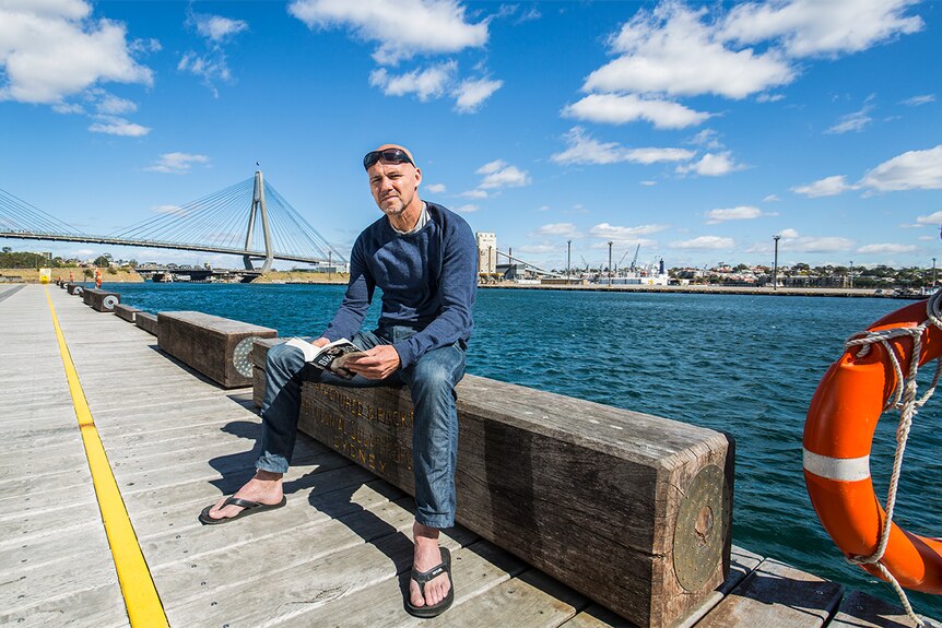 A man sits outside on a boardwalk with Sydney's ANZAC bridge in the background