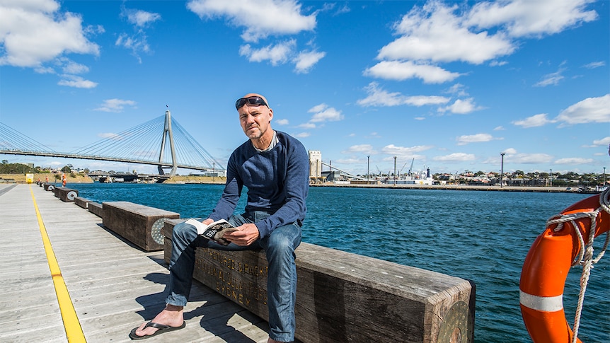 A man sits outside on a boardwalk with Sydney's ANZAC bridge in the background