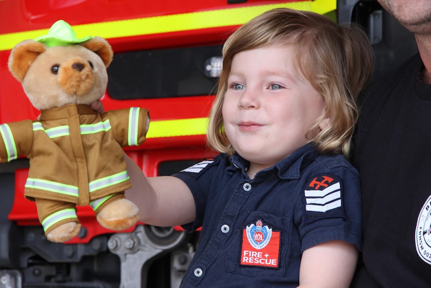 4-year-old boy with long blonde hair and blue eyes holding teddy in one hand in front of firetruck