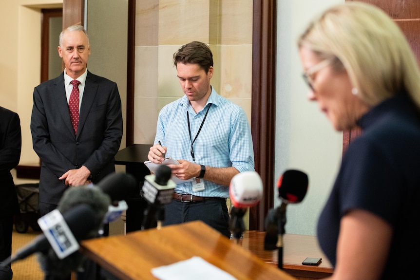 Three people stand in a room watching a press conference