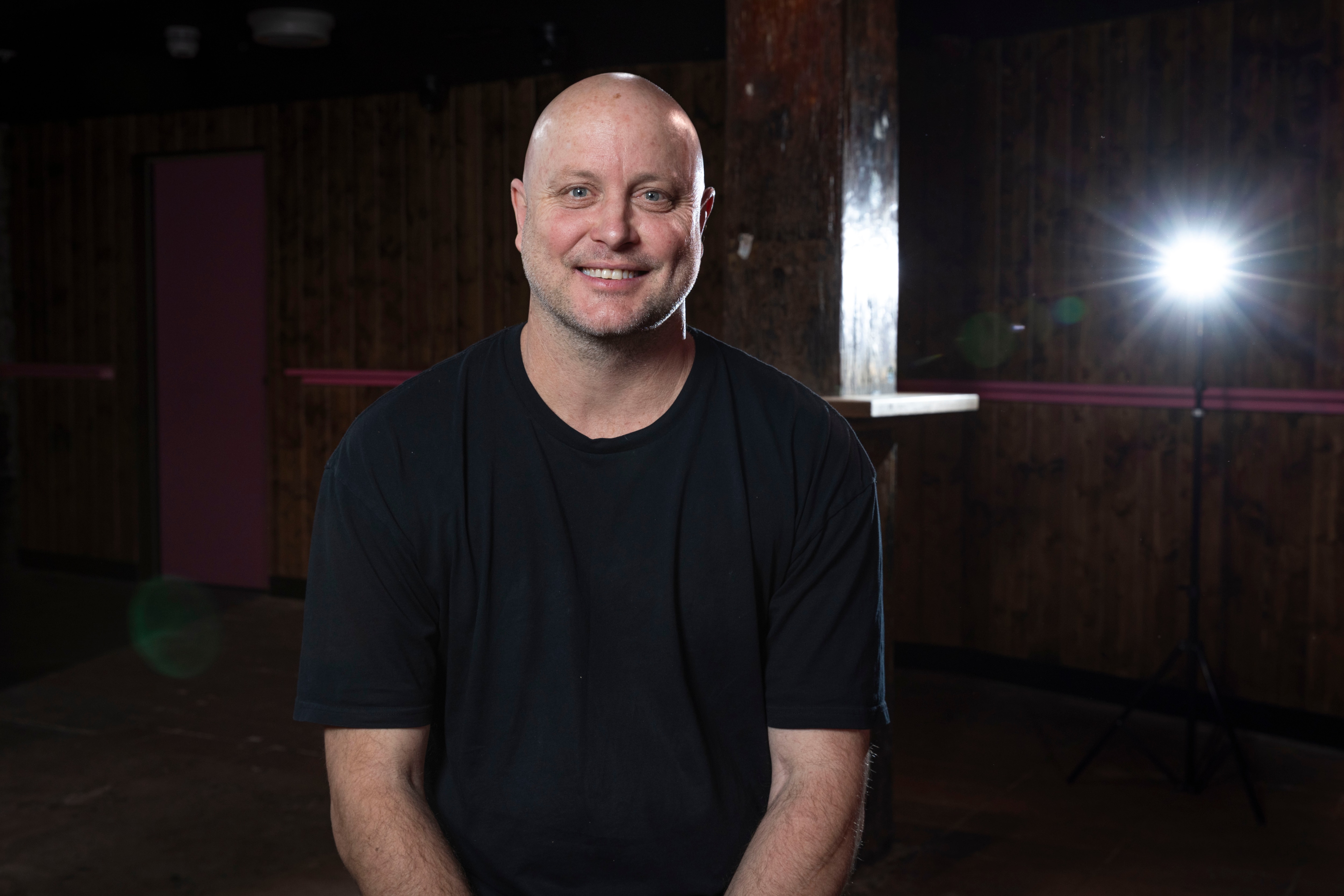 Dane Gorrel sitting and smiling in a black shirt inside a dark room.