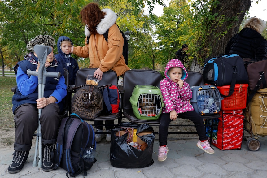 Family sit on bench with pet carriages and belongings as they wait for evacuation buses.