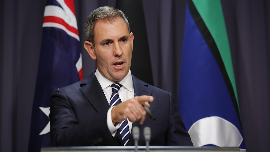 Jim Chalmers, a man in a dark suit, stands behind two microphones and in front of an Australian flag, pointing