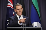 Jim Chalmers, a man in a dark suit, stands behind two microphones and in front of an Australian flag, pointing