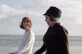 Two women in 1920s costumes walk side by side along a beach, chatting and smiling.