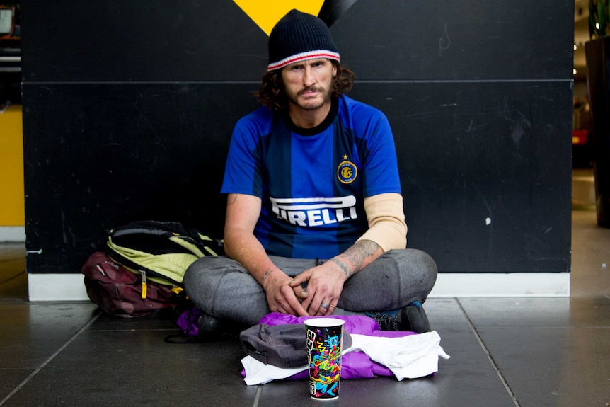 A bearded man in a beanie sits on a footpath with an empty cup placed in front of him.