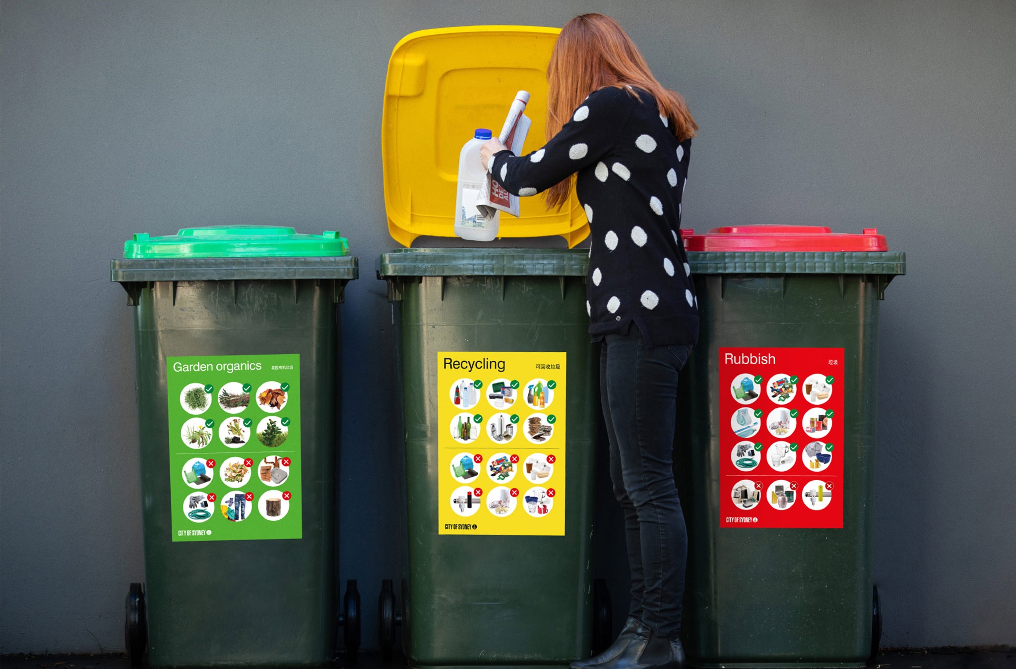 Green, yellow and red bins agianst a dark grey wall, a woman is putting an empty milk bottle into the recycling bin