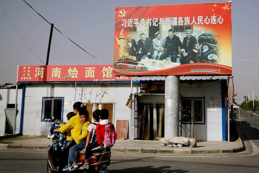 一名妇女骑着摩托车带着几个孩子路过一张中国国家主席习近平的照片