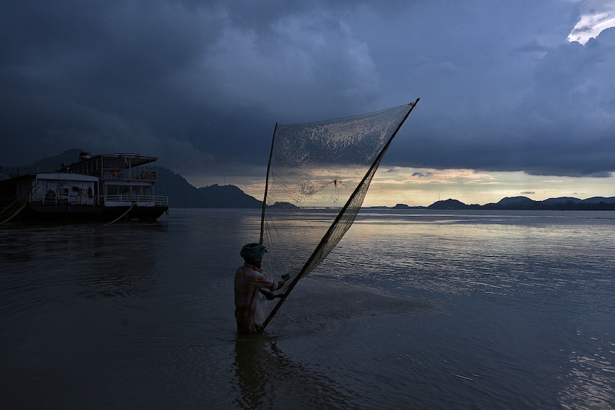 A man in waist deep water on the edge of a rivers casts a large fishing net